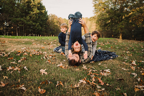 father holds one of his three sons upside down while they play in the grass