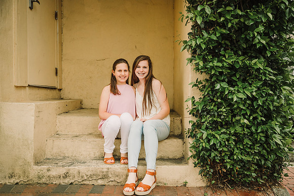 sisters sitting together by a yellow wall and greenery during a family photo session in Marietta, GA
