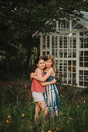 sisters hug each while standing in a flower field in front of a greenhouse