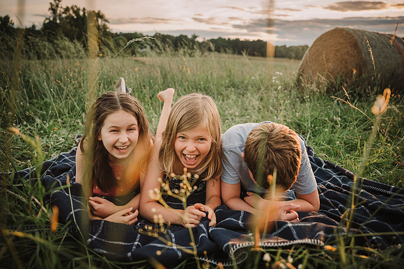 three siblings laugh together while laying in a field of flowers by monica carlson