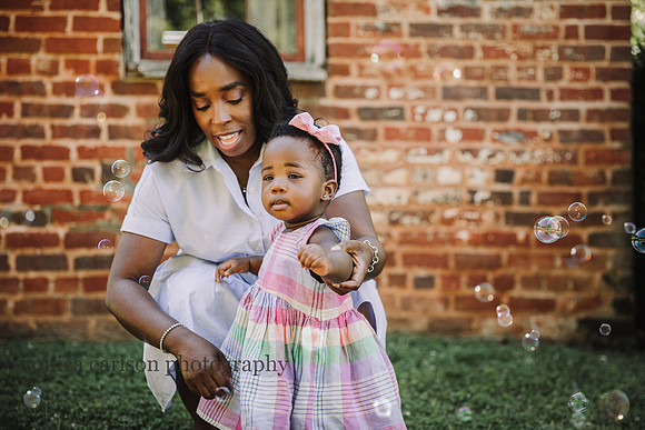 mom steadies a one year old girl who is looking at bubbles in front of a brick building