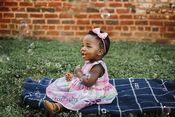 happy african american baby girl plays with bubbles while sitting on a blanket during a family photo session in roswell, GA