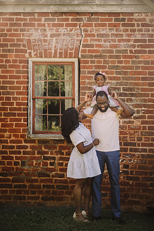 little girl happy sits on her fathers shoulder while her mother looks up adoringly while standing in front of a brick building