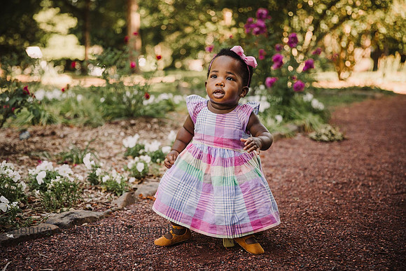 chubby one year old wearing a pretty pastel party dress sits on a stool in a garden
