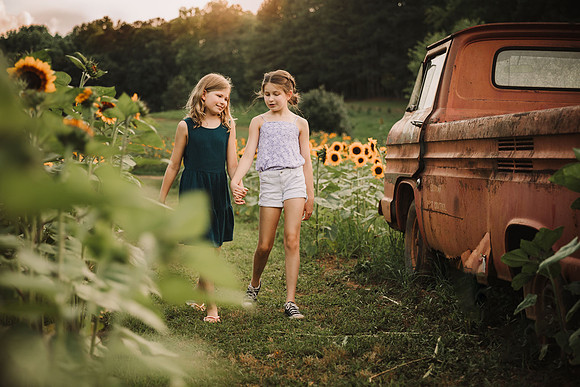 sisters talking as they walk through a field of flowers and past a old red truck by monica carlson photography