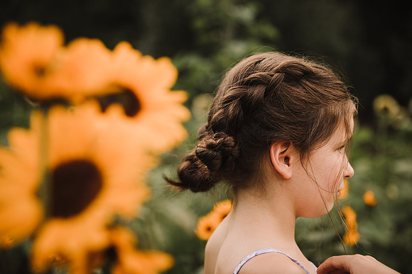 a brunette girl brushing a whisp of hair away from her face while standing on a field about sunflowers