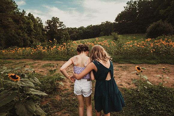 two girls walking away from the camera and towrds a field of sunflowers with their arms wrapped around each other