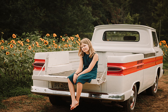 blonde little girl smiling and sitting in a pick up truck that is parked in front of sunflower field