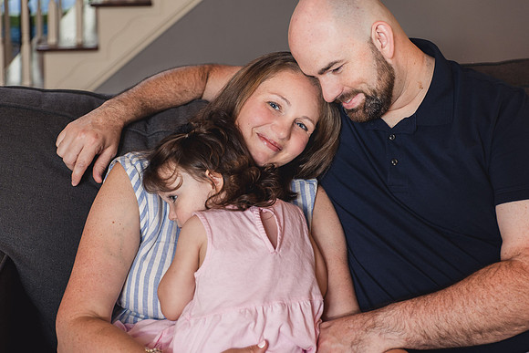 husband and wife cuddle their preschool aged daughter on the couch of their home during a photo session with monica carlson