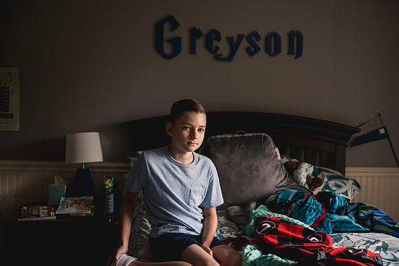 serious 5th grade year old boy sitting on his bed and his name is on the wall behind hi,