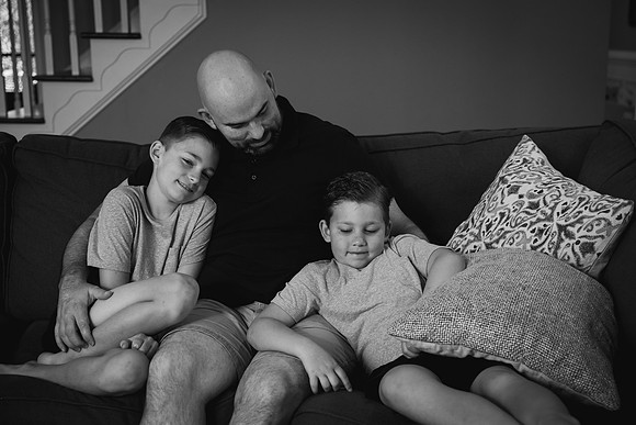black and white image of two elementary aged boys cuddling with their dad on the couch during a home photo session