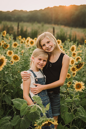 two smiling sisters stand in a field of sunflwowers dunring a family photo session with monica carlson