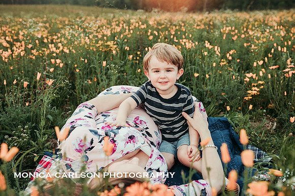 young boy smiles at the camera while his mother lays on a blanket in a flower field during a maternity photo session