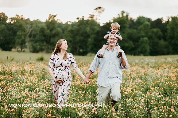little boy sits on his father shoulders while he and his pregnant wife walk through a wild flower field by monica carlson photography