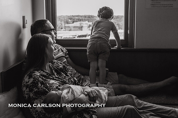 toddler boy looks out hospital window while parents watch and hold newborn