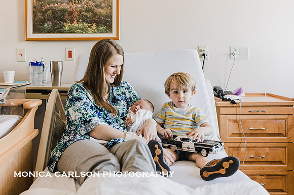 mother in her hospital bed with newborn and toddler son while the older one plays with a toy car