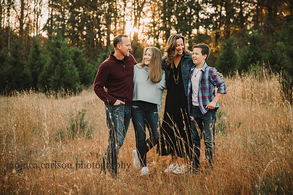 family of four stands among tall grass with christmas trees in the background at sunset