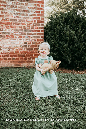 Happy one year old sitting on a stool with a brick building behind her and she is clutching a giraffe toy