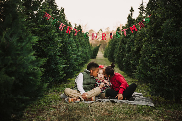todder sits between her sibilings while they kiss her cheeks. They are sitting below a merry christmas sign at Bottoms Christmas tree Farm