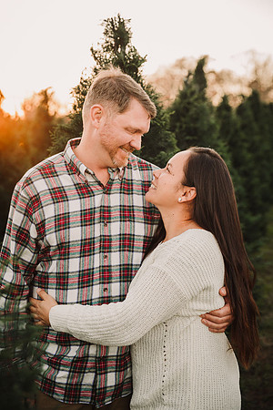 husband and wife look at each other lovingly while at a CHristmas tree farm at sunset