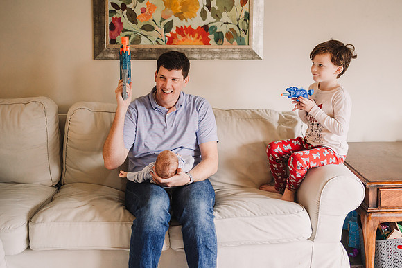 dad plays with son while holding newborn