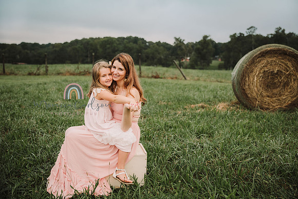 mom and daughter sit on a antique block on a farm field with a hay of bale behind them
