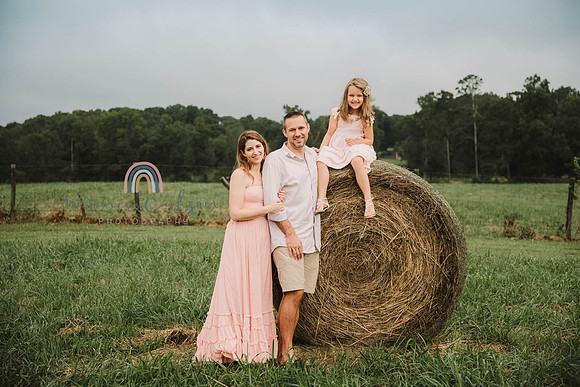 husband and wif stand next to a hay bale and their girl sits on a hay bale