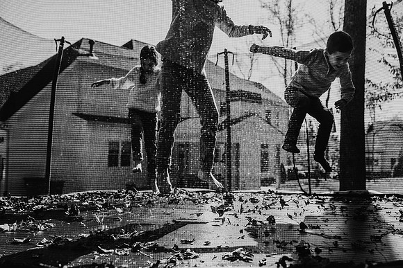 black and white photo of three siblings jumping on a trampoline behind their house by monica carlson