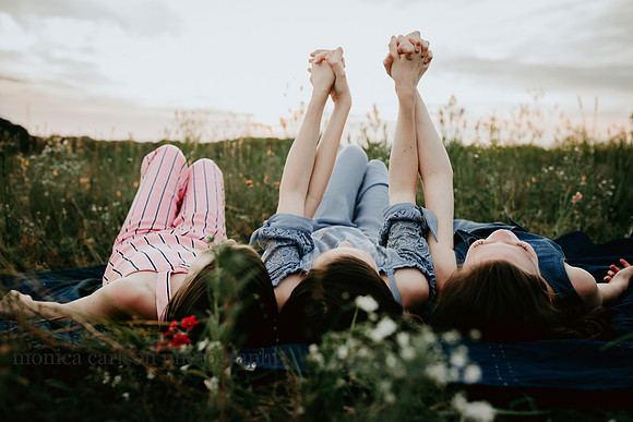 three sisters laying in a wildflower field holding hands in the air by Monica carlson photography