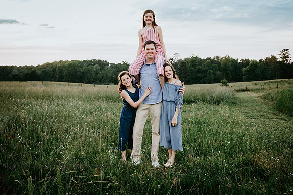 father with his three daughters and one of them is on his shoulders while they are standing in a open field
