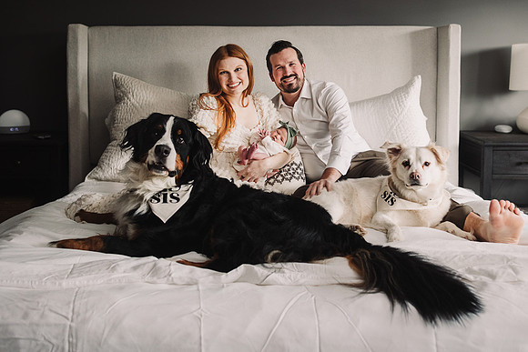 Newborn being held by her mom while sitting in a bed with dad and two dogs