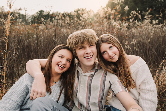 three teen/tween siblings laughing together while sitting in a field