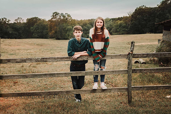 teen boy and girl leaning and standing on a wooden fence in front a ope field