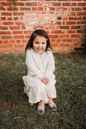 adorable little girl sitting on a stool with a brick building behind her