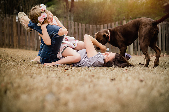 woman laying on the ground while a child grabs her leg and dog kisses her face