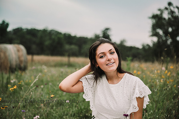 young woman brushes the wind blown hair from her face while sitting in a flower field.