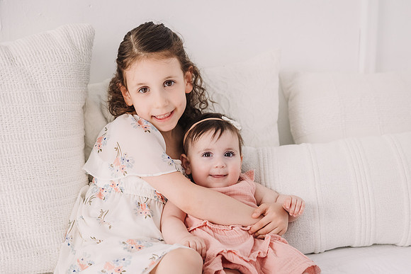 Dark haired sisters cuddle while sitting on a bed