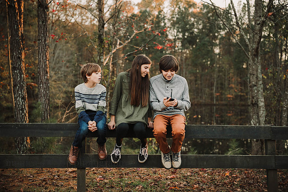 three siblings sitting on a fence and looking at a phone